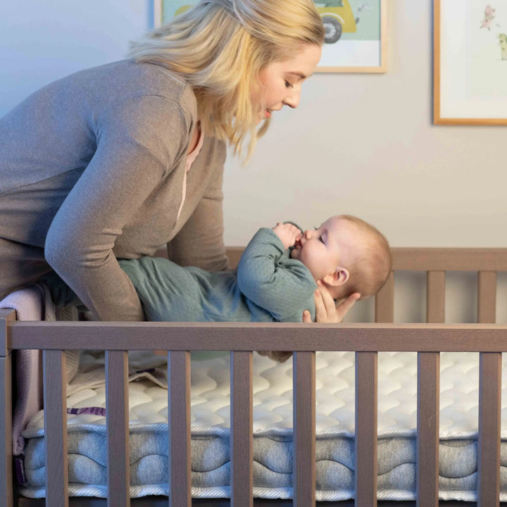 ClevaMama Premium Deluxe ClevaFoam Pocket Sprung Mattress — parent placing baby onto breathable, supportive mattress with ClevaFoam technology and individual pocket springs, designed to provide ultimate comfort, airflow, and safe sleep support in a modern wooden cot.