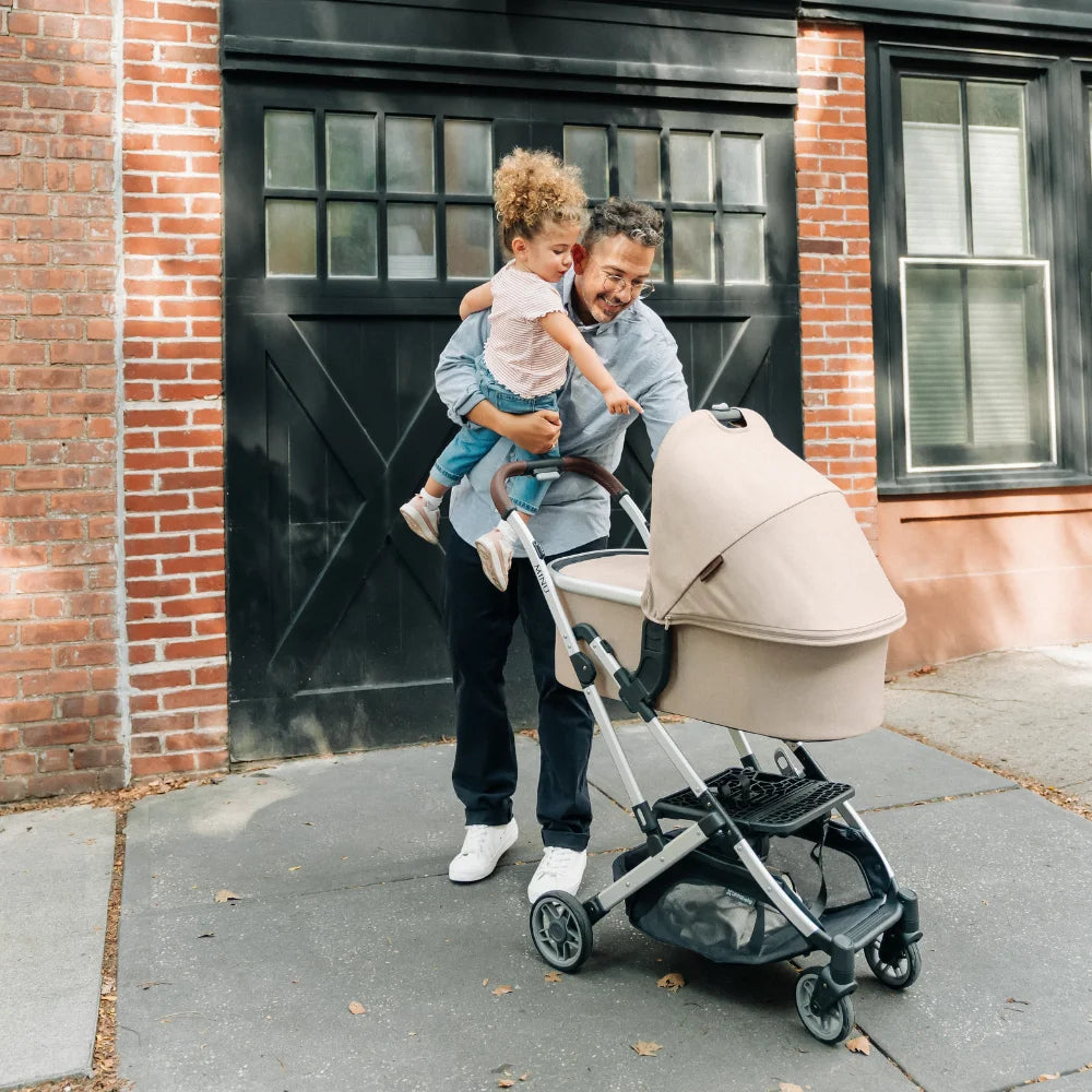 UPPAbaby Carrycot V2 in Declan, an oat melange fabric on a silver frame on the Minu V2 stroller frame with the use of adapters. A father is holding his toddler on his hip, that is pointing in to the baby that is asleep in the carrycot. They are walking down a footpath.