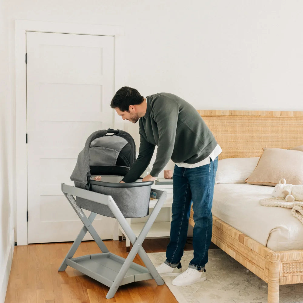This image is of a parent cooing at their child in the UPPAbaby Carrycot V2 in Greyson which is a Grey melange fabric with a carbon frame, on a light grey carrycot stand. It is placed in a bedroom, a few feet from the bed.