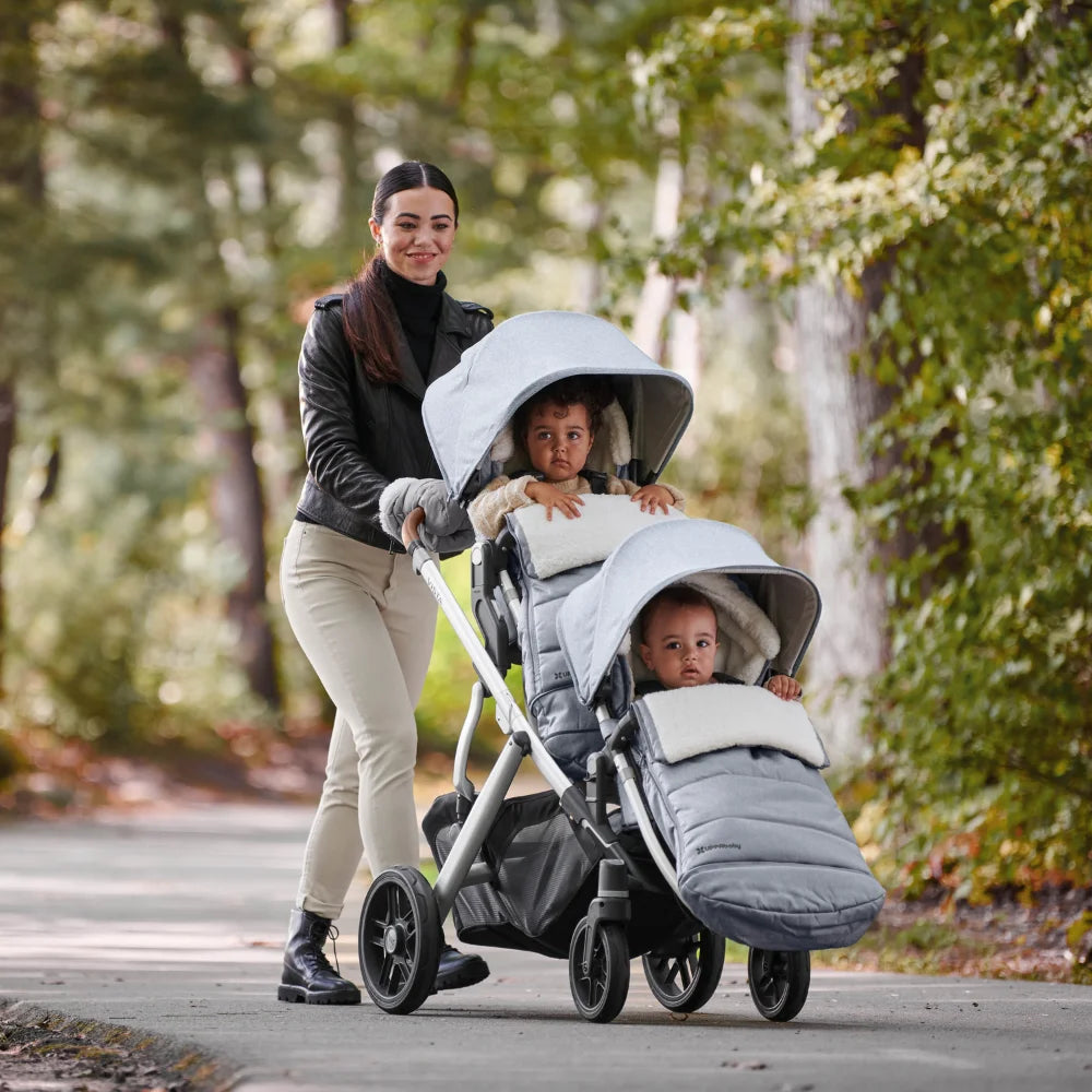 This image shows a mum pushing her two kids in the double configuration of the Vista with a toddler seat and a RumbleSeat on the bottom. The cosy ganoosh footmuff is on each one and the mum has her hands in the handmuffs.