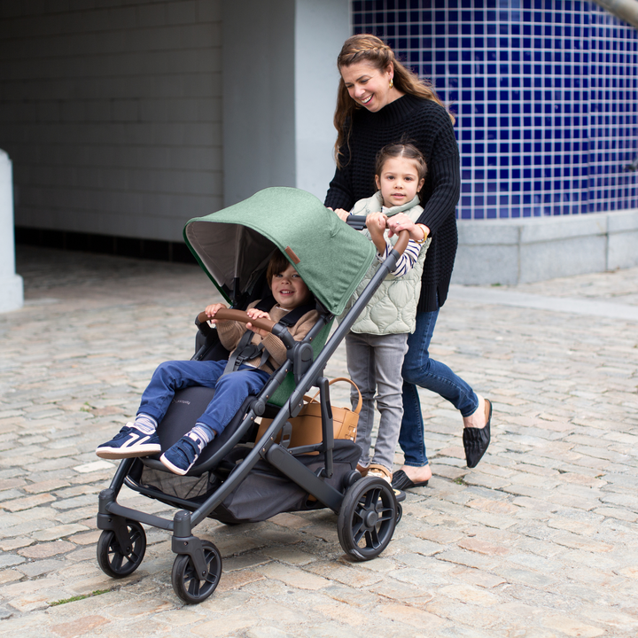 A Mum pushing her Cruz stroller in the Gwen fashion with her younger child smiling in the main seat unit and the older child standing on the piggyback buggy board.