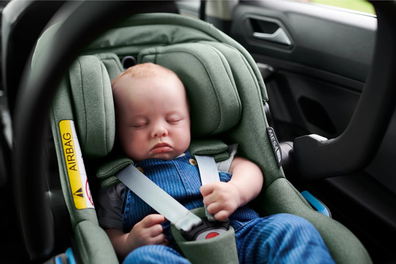 UPPAbaby Mesa i-Size Infant Car Seat in the green Gwen or Emmet fashion safely installed in the back seat of a car.  A small baby is sleeping soundly while securely fastened in the car seat.
