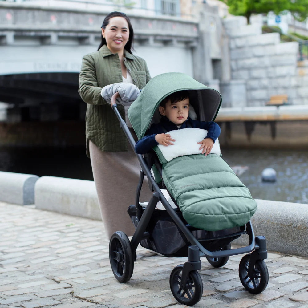 This image depicts a mother pushing her child in the UPPAbaby Vista V2 stroller in the green gwen fashion along a river on a cobbled path. The child is in the stroller with the footmuff attached and the mother is using the CozyHandmuffs.
