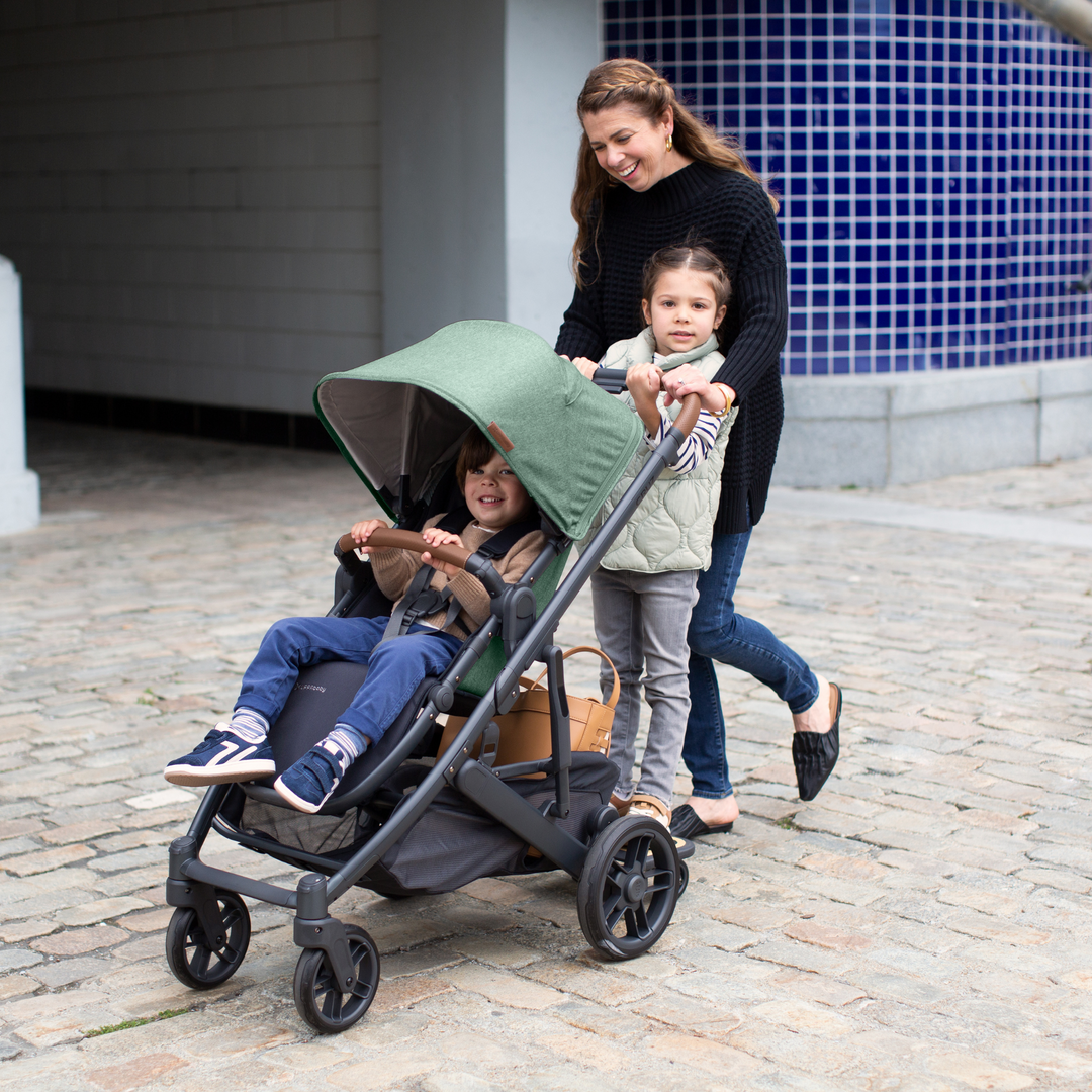 UPPAbaby Cruz V2 in the Gwen fashion which is a green melange fabric on a carbon chassis with saddle REACH certified leather accents. A smiling mother is pushing her toddler in the toddler seat of the stroller, who is smiling widely and her older daughter is standing on the piggyback board looking into the camera. The large basket is full of bits and bobs.