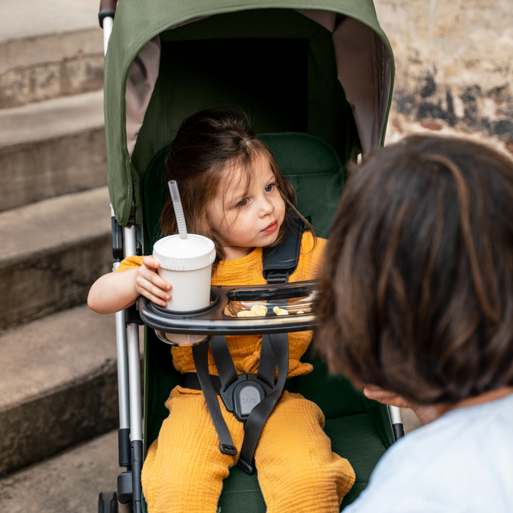 UPPAbaby Minu V2 lightweight travel stroller in Emelia which is a green fabric with chestnut REACH certified leather accents on a silver frame. This image shows a toddler bucked safely into the stroller in a bright yellow jumpsuit using the Minu snack tray accessory. She has a cup with a straw for her drink and a few crackers in the tray.