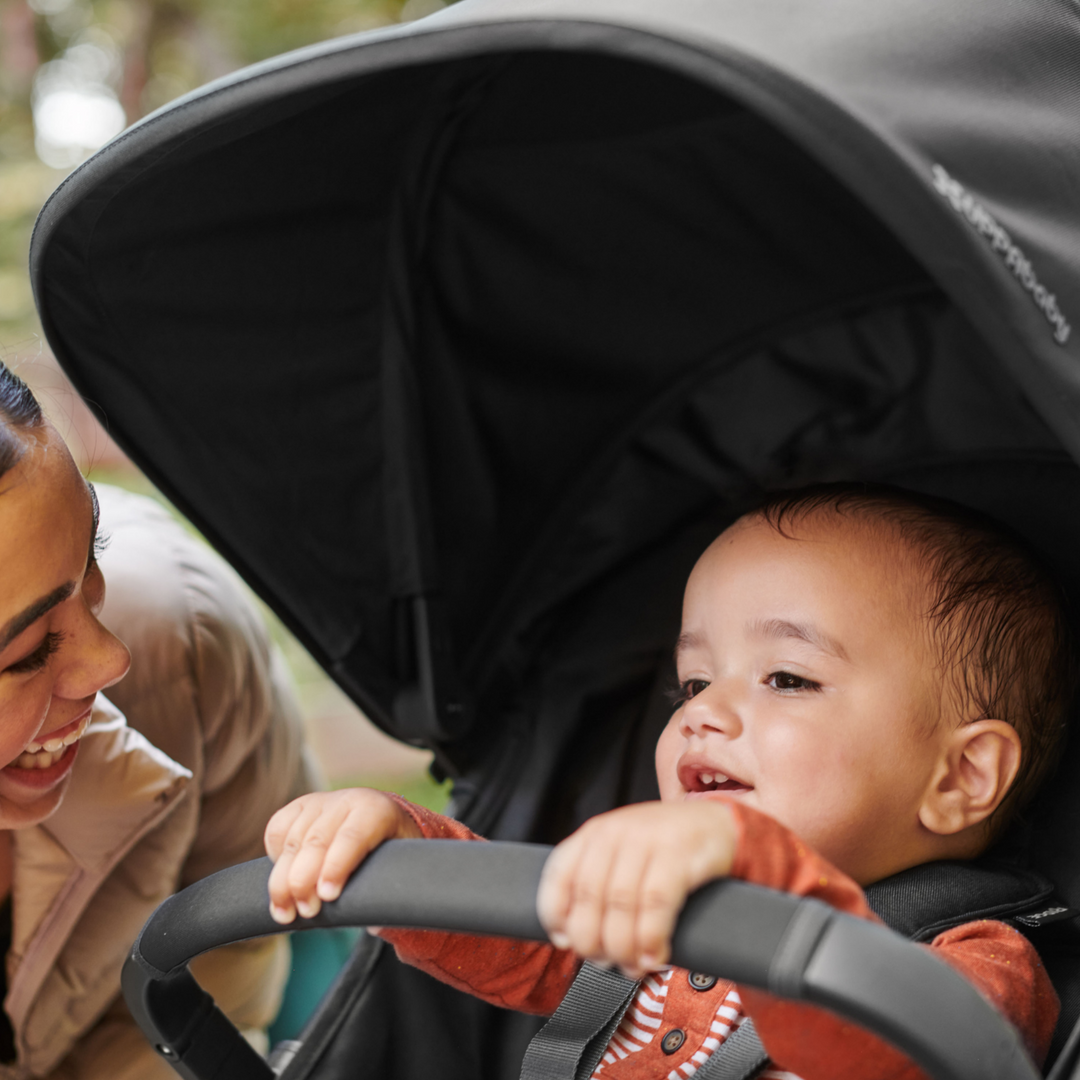This is a lifestyle image of a Mum smiling at her child holding on to the bumper bar as he is strapped into the stroller on their walk