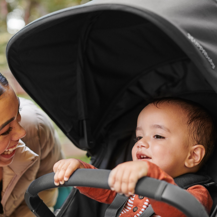 This is a lifestyle image of a Mum smiling at her child holding on to the bumper bar as he is strapped into the stroller on their walk