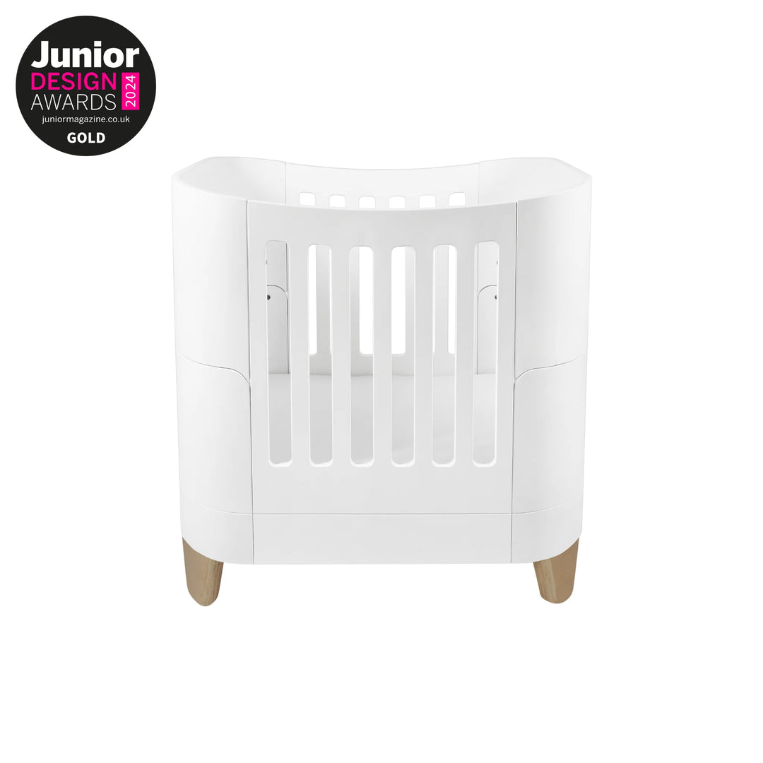 Serena Mini Cot with natural wooden legs in a modern white curved design, award-winning Gold at the Junior Design Awards, premium space-saving baby cot for newborns and small nursery spaces.
