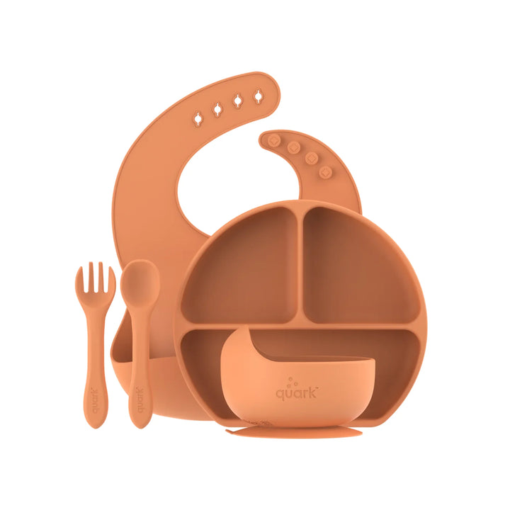 Orange silicone baby dining set including an adjustable bib, divided suction plate, bowl, fork, and spoon arranged on a white background with Quark branding visible on the utensils and bowl.