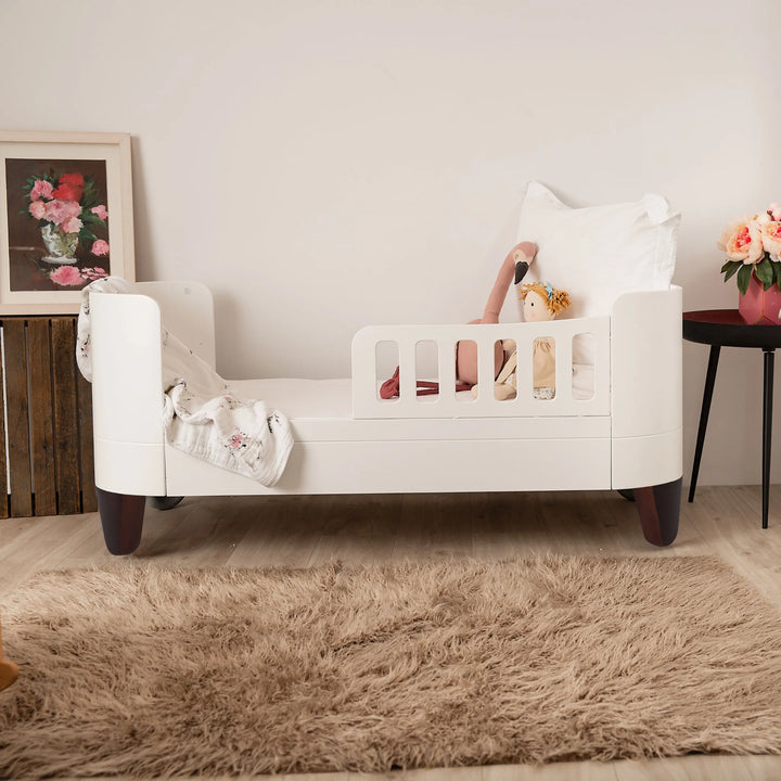 Gaia Baby Serena First Bed with toddler rail and walnut legs styled in a modern toddler bedroom, featuring the brand’s curved white design, soft bedding, plush toys and warm neutral décor to showcase a safe and stylish transition bed.