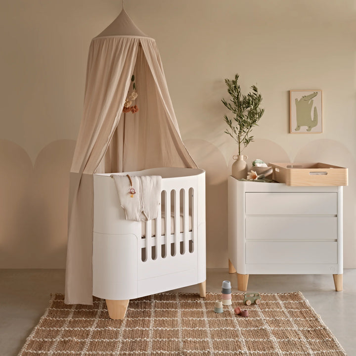 Serena Mini Cot with natural wooden legs styled in a modern neutral nursery with matching white dresser, Maia canopy, Hera changer top and Scandinavian décor, showcasing a premium space-saving sleep setup for babies.