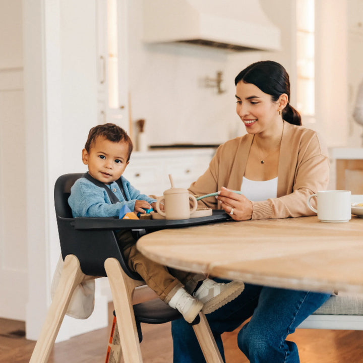Parent feeding toddler seated in UPPAbaby Ciro High Chair Jake in modern kitchen, showcasing ergonomic design, adjustable tray, and stylish natural wood legs—premium baby high chair for mealtime.