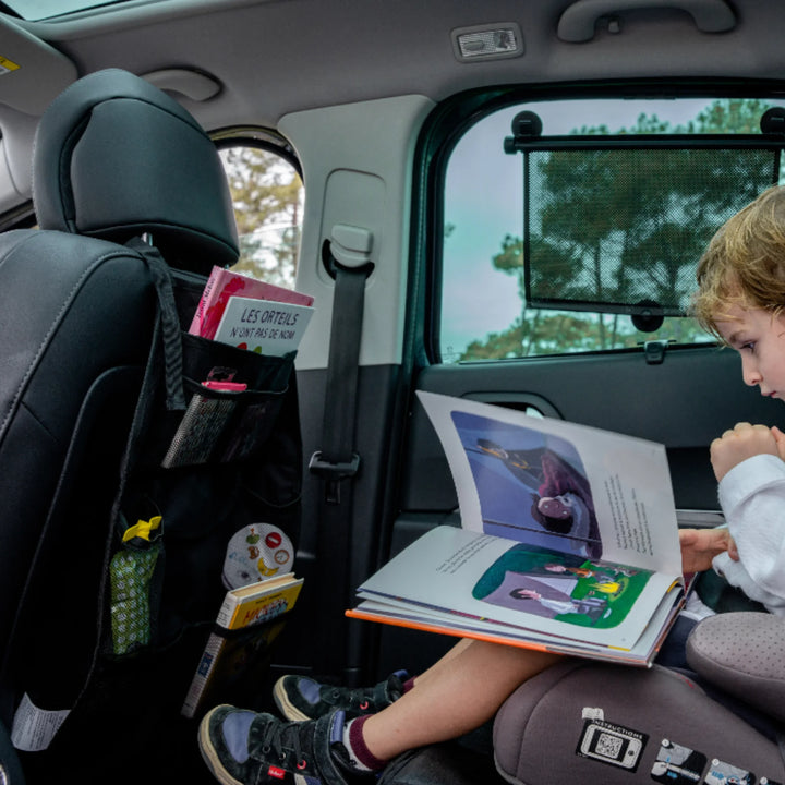 EziMoov EZI Travel Plus car seat back organiser installed on a front seat, stocked with children’s books, snacks, and travel essentials. A child in a car seat reads a picture book beside it, demonstrating how the organiser keeps kids’ items tidy and accessible during family road trips and everyday car travel.