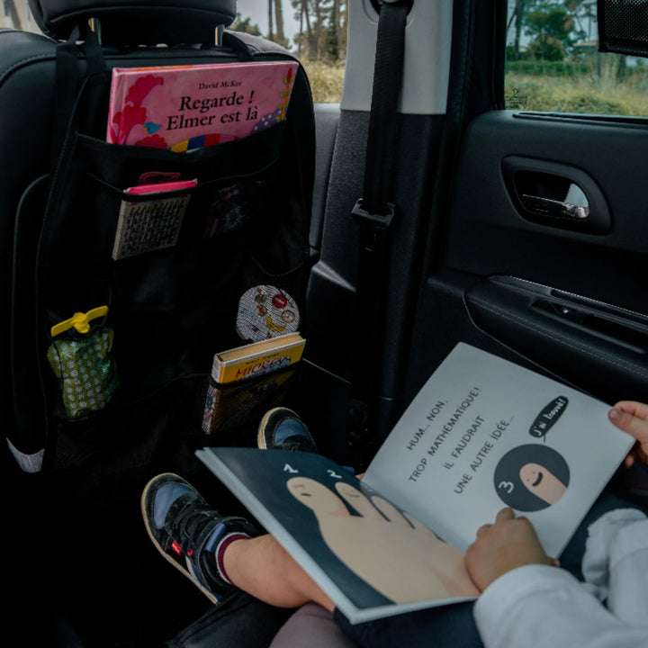 EziMoov EZI Travel Plus car seat back organiser attached to the front seat, stocked with children’s books, snacks and travel essentials. A child in the back seat reads a picture book beside it, demonstrating how the organiser keeps kids’ items tidy, organised and easy to reach during family car trips and everyday travel.