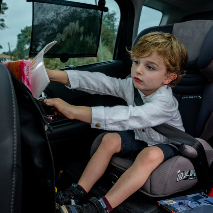 Young child seated in a car seat reaching into the EziMoov EZI Travel Plus car seat back organiser to retrieve a book. The organiser is attached to the front seat and holds children’s books, toys, and travel essentials, demonstrating how it keeps items organised, easy to reach, and ideal for family road trips and everyday car travel.