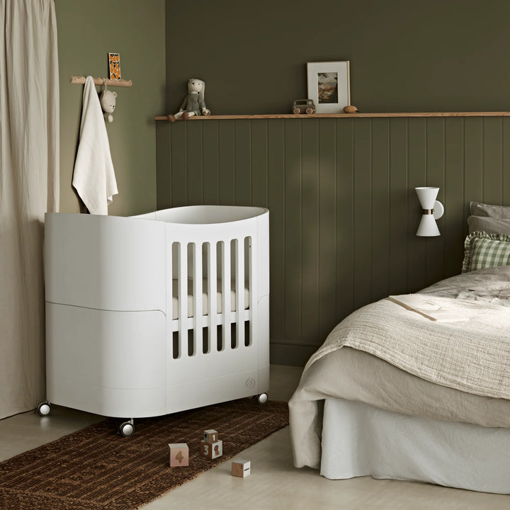 Serena Mini Cot on wheels positioned beside a parent’s bed in a modern, earthy-toned bedroom, showcasing its compact curved design, easy mobility and stylish Scandinavian aesthetic for newborn sleep.