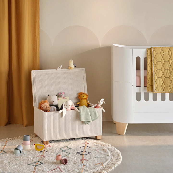 Gaia Baby Serena Mini Cot in white with natural legs styled in a modern nursery beside an ecru Serena footstool toy box filled with soft toys, set against mustard curtains and a neutral decorative rug for a contemporary Scandinavian baby room look.