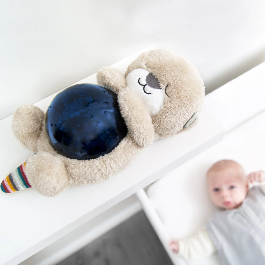 ZAZU Otto the Otter ocean projector night light plush on a bedside table beside a baby.