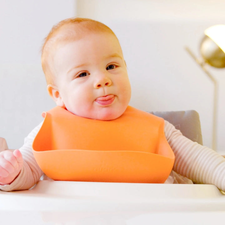 Smiling baby wearing Feedi by Quark silicone bib in terracotta orange, BPA-free waterproof feeding bib designed for easy cleaning and mess-free baby-led weaning.