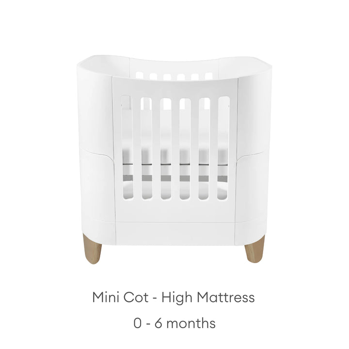 “Serena Mini Cot with natural wooden legs shown in the high-mattress position for newborns aged 0–6 months, featuring a modern white curved design and breathable slatted sides.