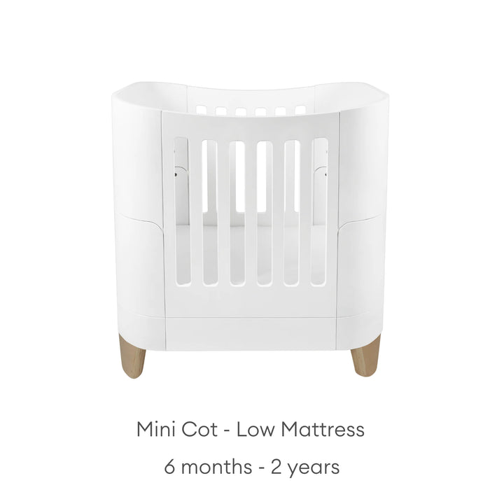 Serena Mini Cot with natural wooden legs shown in the low-mattress position for babies aged 6 months to 2 years, featuring a modern white curved design and safe breathable slatted sides.