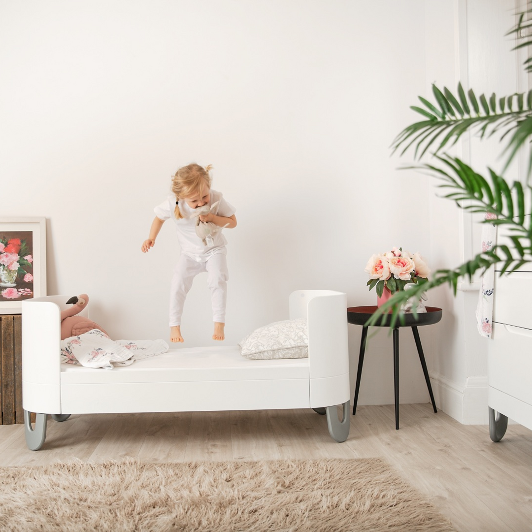 Serena Cot Bed All White as First bed. Lifestyle image showing little girl jumping on the bed