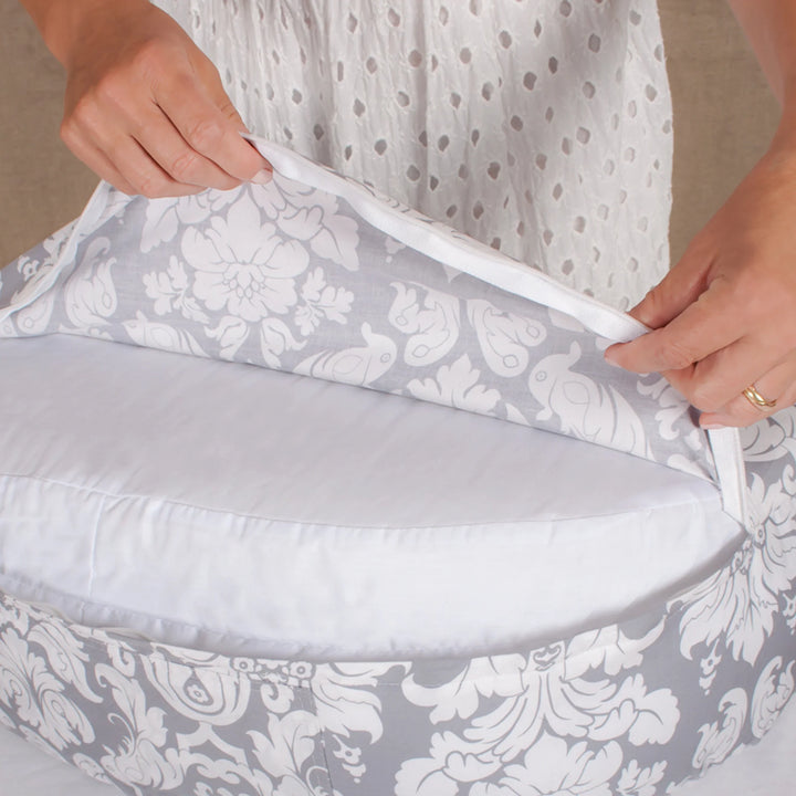 Close-up lifestyle image of a parent unzipping the Bebe au Lait Complete Nursing Pillow in Chateau Silver, revealing the removable washable insert beneath the grey and white damask cover, highlighting easy-care design and premium breastfeeding support.