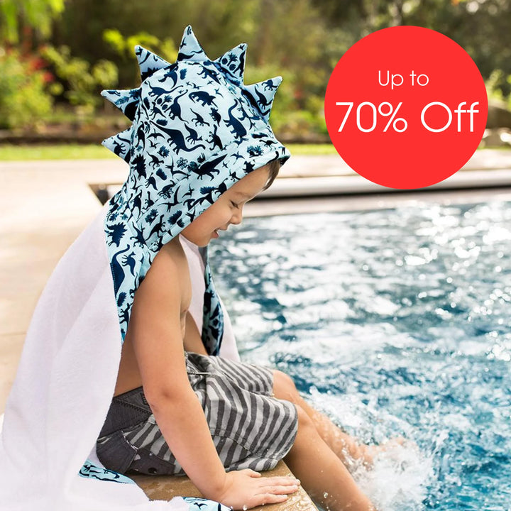 Child sitting by a swimming pool wearing a Bebe Au Lait dinosaur-print hooded towel with 3D spikes, splashing feet in the water, featured in a summer sale promotion with ‘Up to 70% Off’.