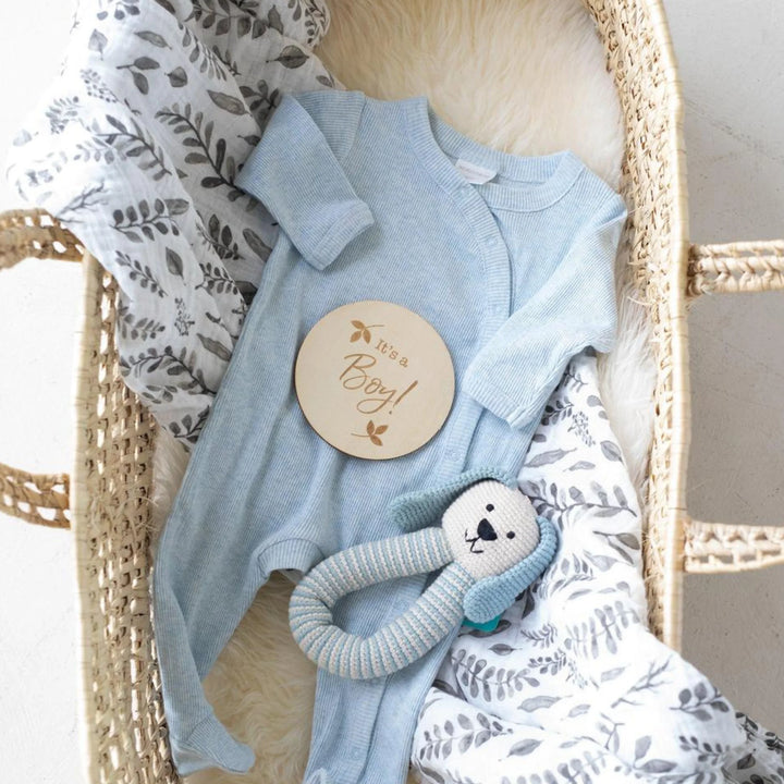 Baby boy announcement flat lay featuring a wooden ‘It’s a Boy!’ milestone disc, blue newborn onesie, knitted dog rattle toy, and leaf-print swaddle in a woven Moses basket, newborn gender reveal photography prop.
