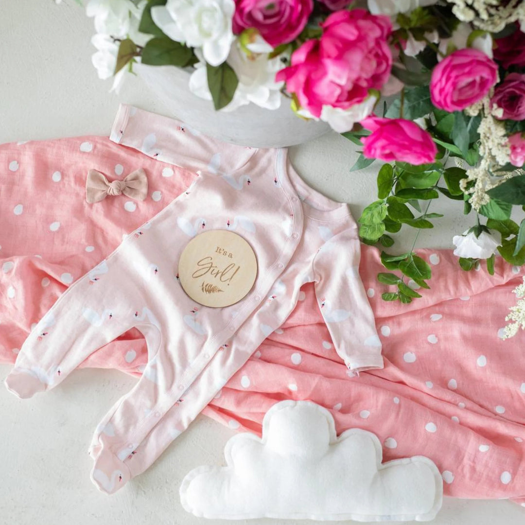 Baby girl flat lay with pink polka-dot swaddle, pink newborn onesie, bow headband, white cloud pillow, and wooden ‘It’s a Girl!’ milestone announcement disc surrounded by bright floral arrangement, newborn gender reveal photography prop.