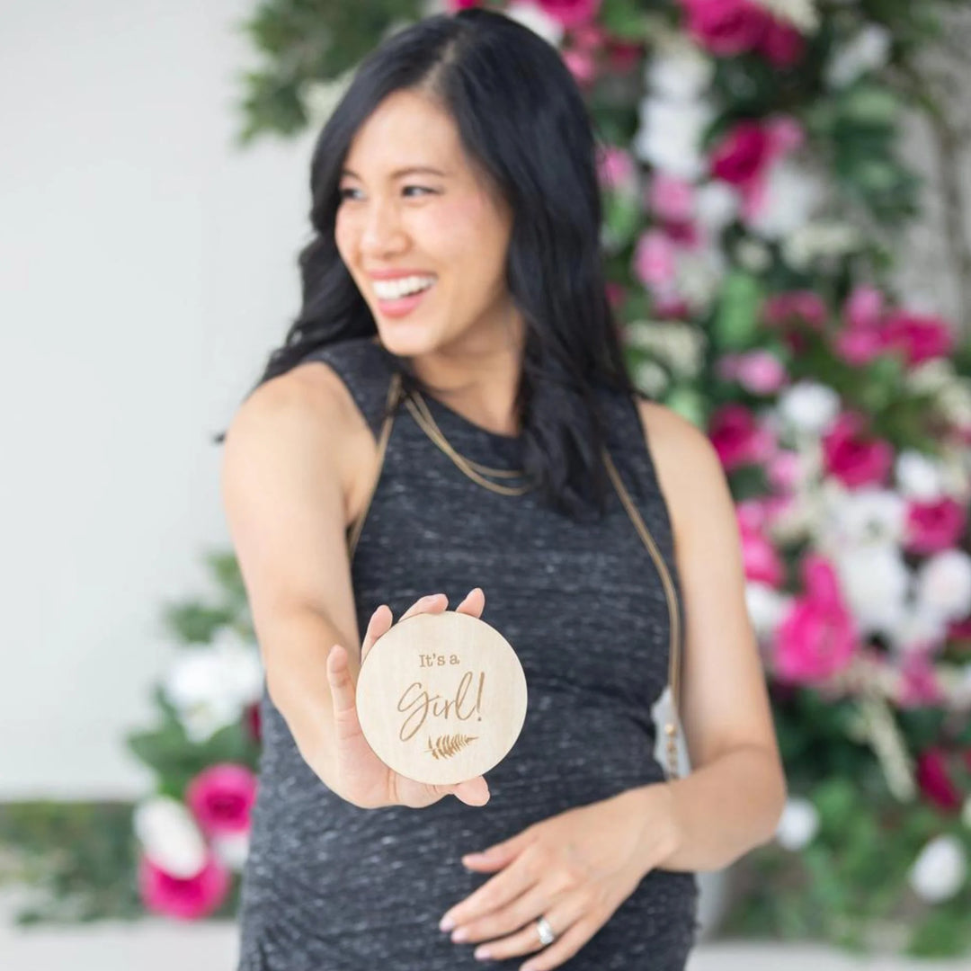 Smiling pregnant woman holding a wooden ‘It’s a Girl!’ milestone announcement disc in front of a bright floral backdrop, gender reveal and maternity photoshoot prop.