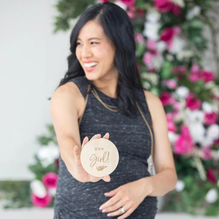 Smiling pregnant woman holding a wooden ‘It’s a Girl!’ milestone announcement disc in front of a bright floral backdrop, gender reveal and maternity photoshoot prop.
