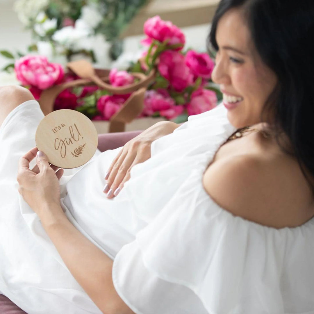 Pregnant woman in a white dress smiling while holding a wooden ‘It’s a Girl!’ milestone announcement disc, surrounded by bright pink flowers, maternity photoshoot gender reveal prop.