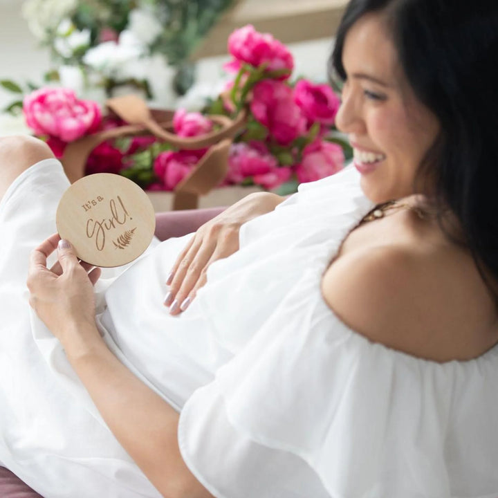 Pregnant woman in a white dress smiling while holding a wooden ‘It’s a Girl!’ milestone announcement disc, surrounded by bright pink flowers, maternity photoshoot gender reveal prop.