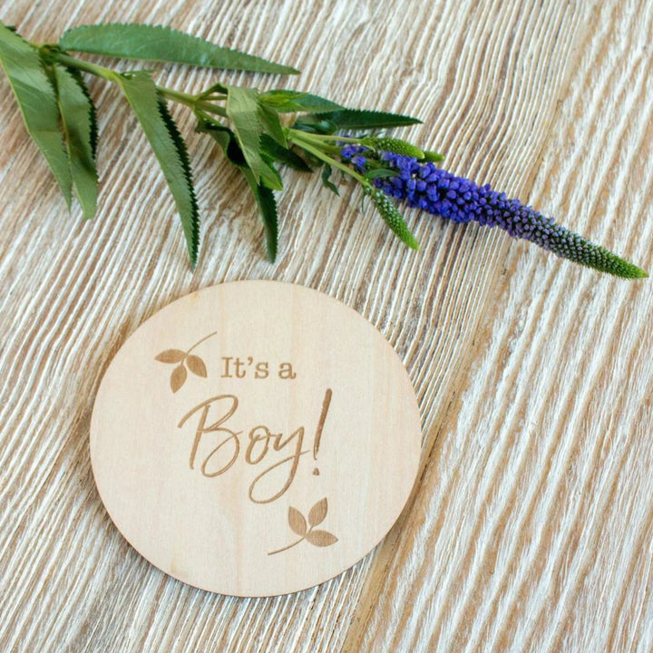 Wooden ‘It’s a Boy!’ baby announcement milestone disc styled on a light wood surface with a purple flower stem, newborn gender reveal photography prop.
