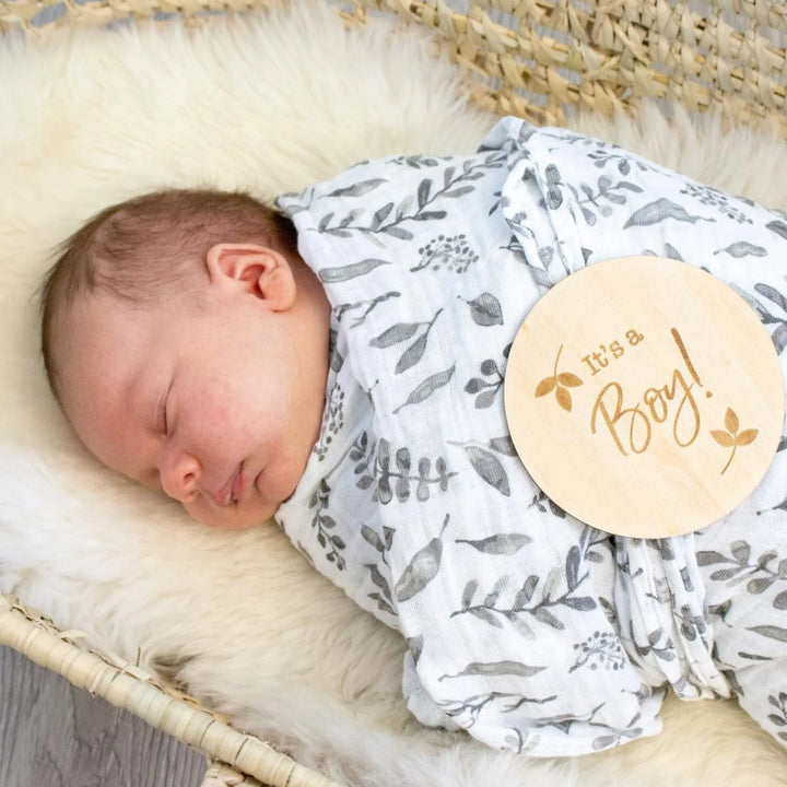 Newborn baby boy sleeping in a leafy swaddle blanket with a wooden ‘It’s a Boy!’ milestone announcement disc on his chest, gender reveal and newborn photography prop.