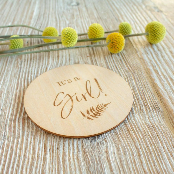 Wooden ‘It’s a Girl!’ milestone announcement disc with engraved fern design styled on a light wood surface beside yellow billy ball flowers, newborn gender reveal photography prop.