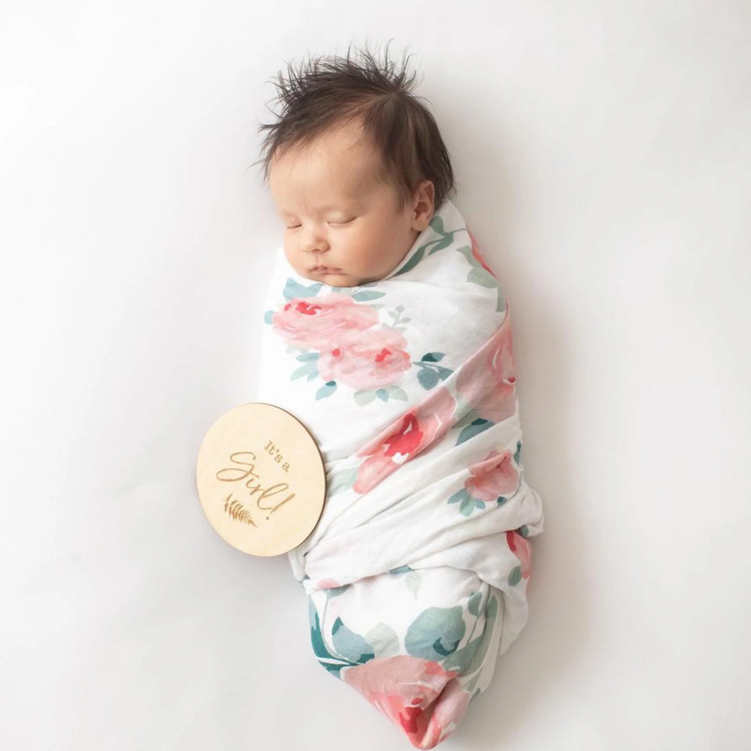 Newborn baby girl wrapped in a floral swaddle blanket, sleeping on a white background with a wooden ‘It’s a Girl!’ milestone announcement disc, newborn gender reveal and birth announcement photography prop.