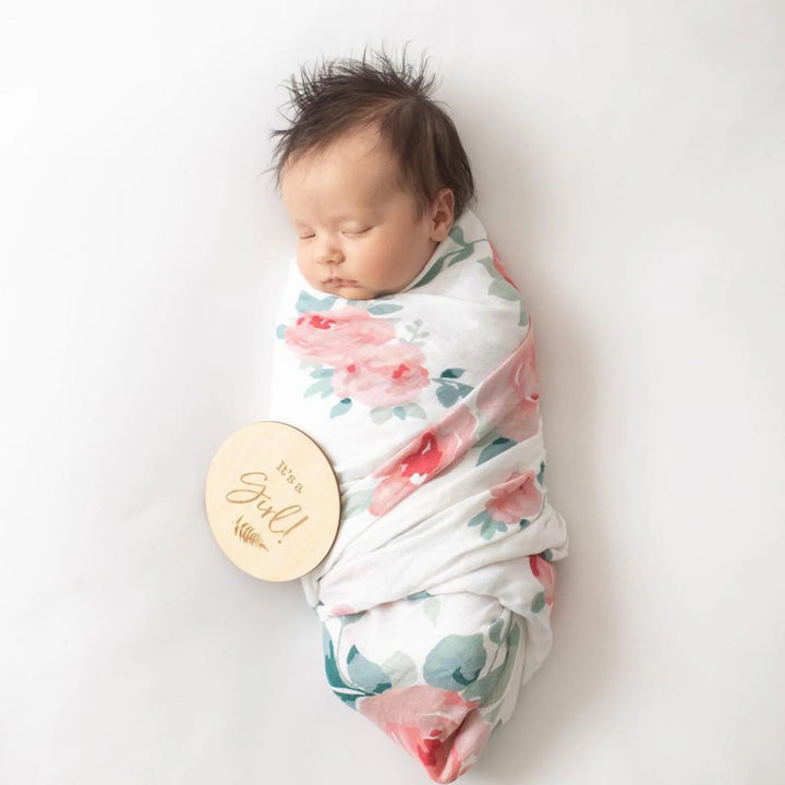 Newborn baby girl wrapped in a floral swaddle blanket, sleeping on a white background with a wooden ‘It’s a Girl!’ milestone announcement disc, newborn gender reveal and birth announcement photography prop.