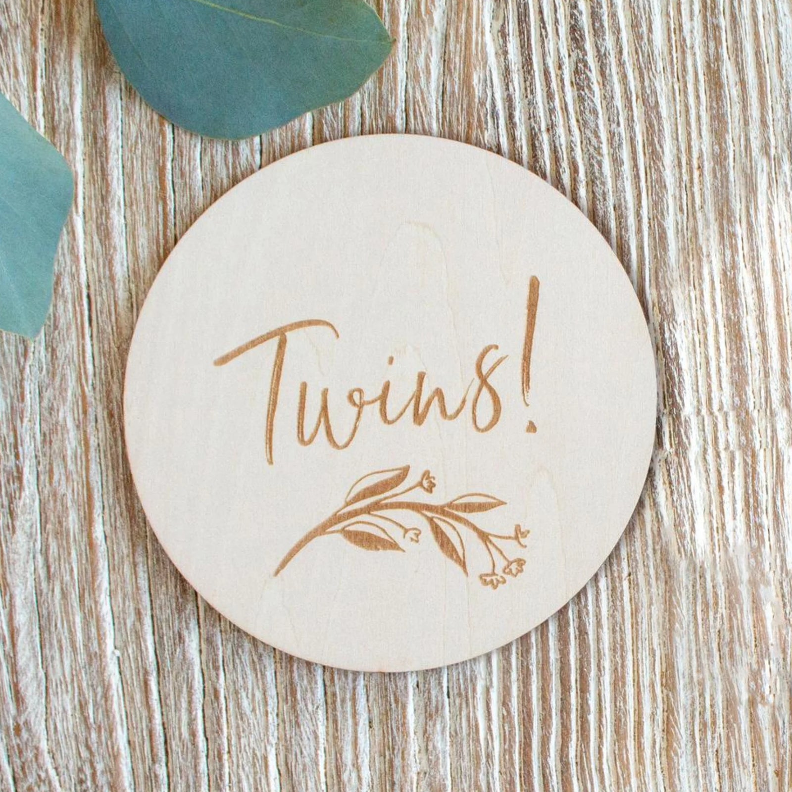 Wooden ‘Twins!’ milestone announcement disc with engraved botanical design, styled on a light wood surface, newborn twin gender reveal and birth announcement photo prop.