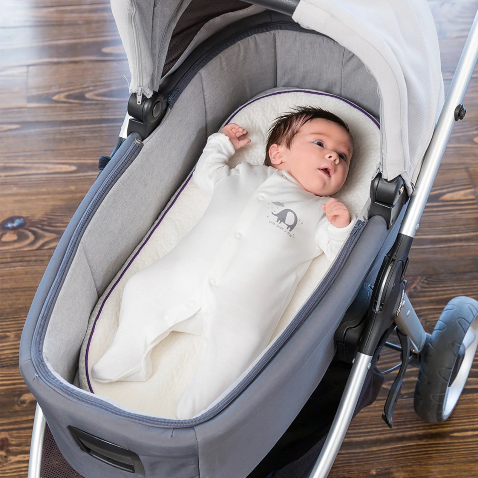 Baby lying in a stroller bassinet on a ClevaFoam Moses Basket Mattress, dressed in a white sleepsuit, looking upward while resting comfortably.