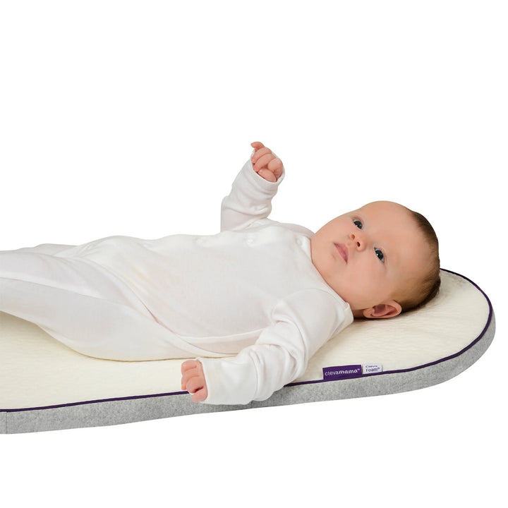 Baby lying on their back on an oval ClevaFoam Moses Basket Mattress, dressed in a white sleepsuit, looking upward with one arm raised