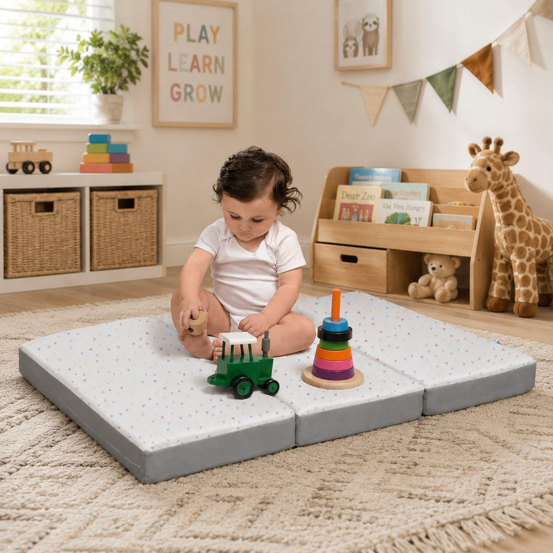 A baby sitting and playing on a foldable 3-in-1 travel cot mattress laid flat as a cushioned play mat in a warm, child-friendly nursery with colourful toys, books, and soft décor.