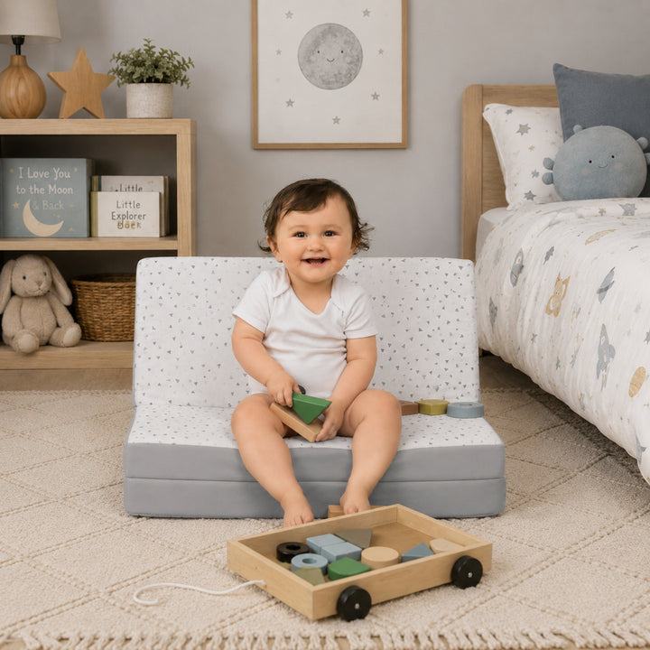 A smiling baby sitting on a foldable 3-in-1 travel cot mattress styled as a mini sofa in a cosy nursery, surrounded by soft toys, books, and wooden play items to showcase versatile comfort and child-friendly functionality.