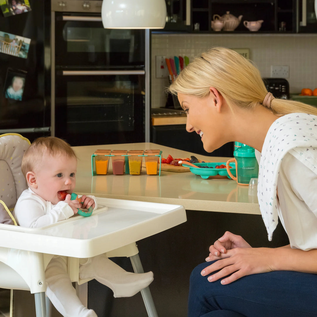 Baby in a high chair using a ClevaMama ClevaFeed fruit feeder while a woman watches and smiles in a kitchen setting.