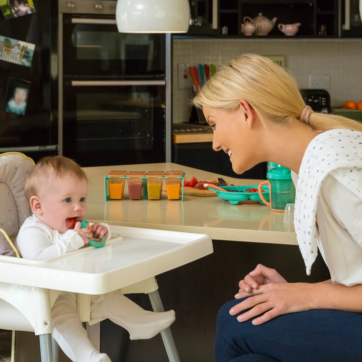 Baby in a high chair using a ClevaMama ClevaFeed fruit feeder while a woman watches and smiles in a kitchen setting.