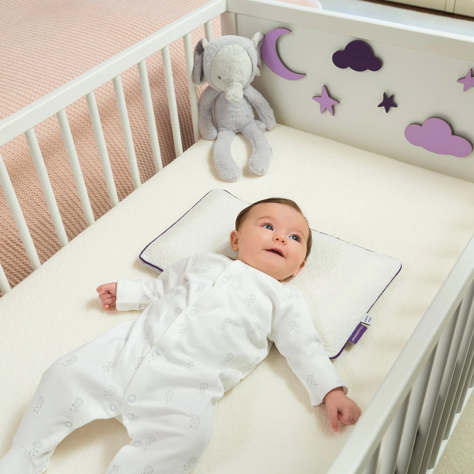 ClevaMama ClevaFoam Baby Pillow in use — baby lying comfortably in cot on breathable memory foam pillow designed to support head and neck alignment, prevent flat head syndrome, and promote safe, restful sleep; soft white cover with purple trim shown in nursery setting.
