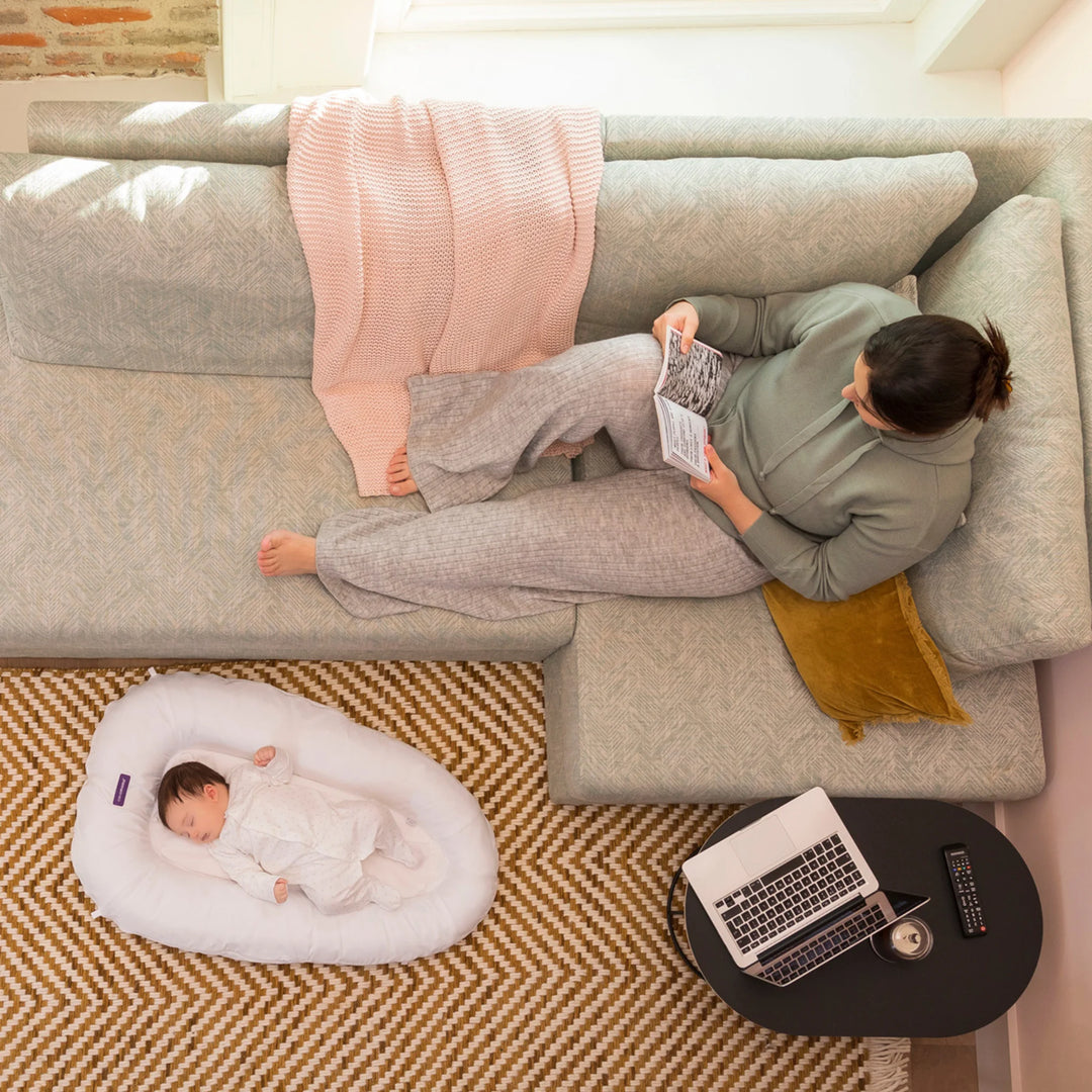 Top-down view of mother relaxing on sofa while baby sleeps in ClevaMama ClevaFoam Baby Pod — premium breathable and supportive baby nest promoting safe, comfortable rest and bonding in a cosy home setting.