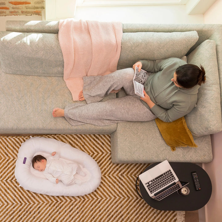 Top-down view of mother relaxing on sofa while baby sleeps in ClevaMama ClevaFoam Baby Pod — premium breathable and supportive baby nest promoting safe, comfortable rest and bonding in a cosy home setting.