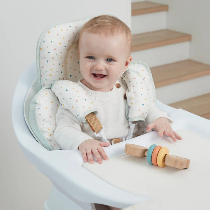 ClevaMama ClevaFoam Head and Neck Support Terrazzo design — smiling baby sitting comfortably in a high chair using ergonomic ClevaFoam support cushion. Breathable and hypoallergenic newborn insert providing head and neck stability to prevent flat head syndrome, ideal for car seats, strollers, bouncers, and feeding chairs.